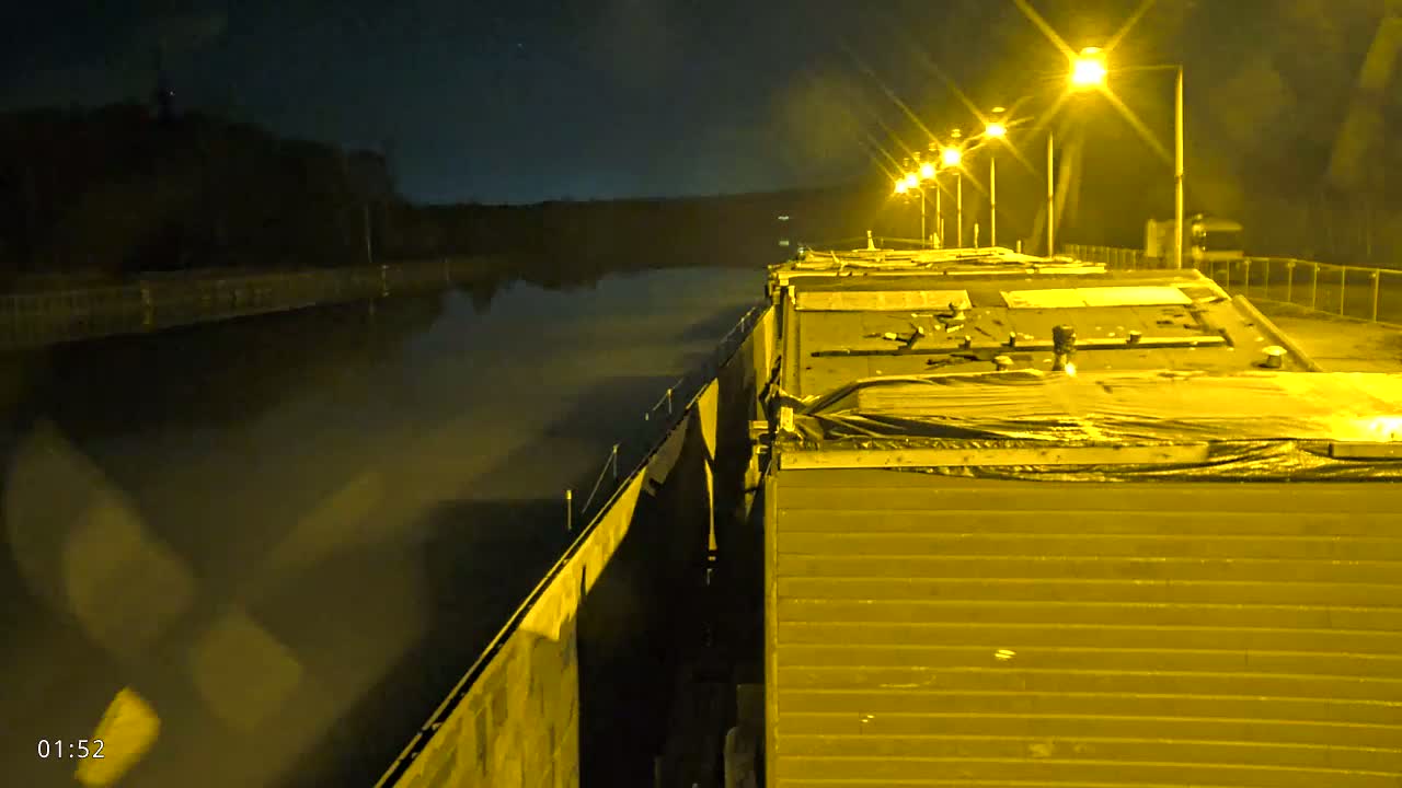 At night, a barge travels down a canal illuminated by bright yellow lights along the bank.