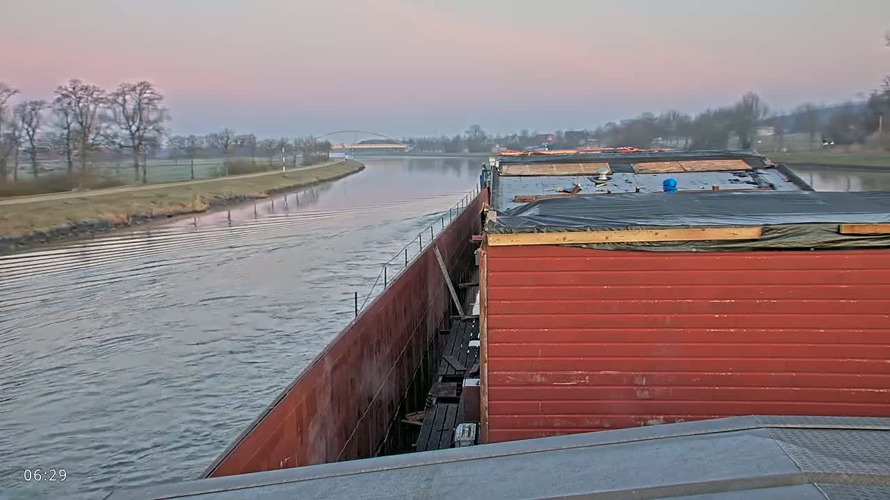 A barge travels down a calm canal at dawn, passing under a bridge in the distance.