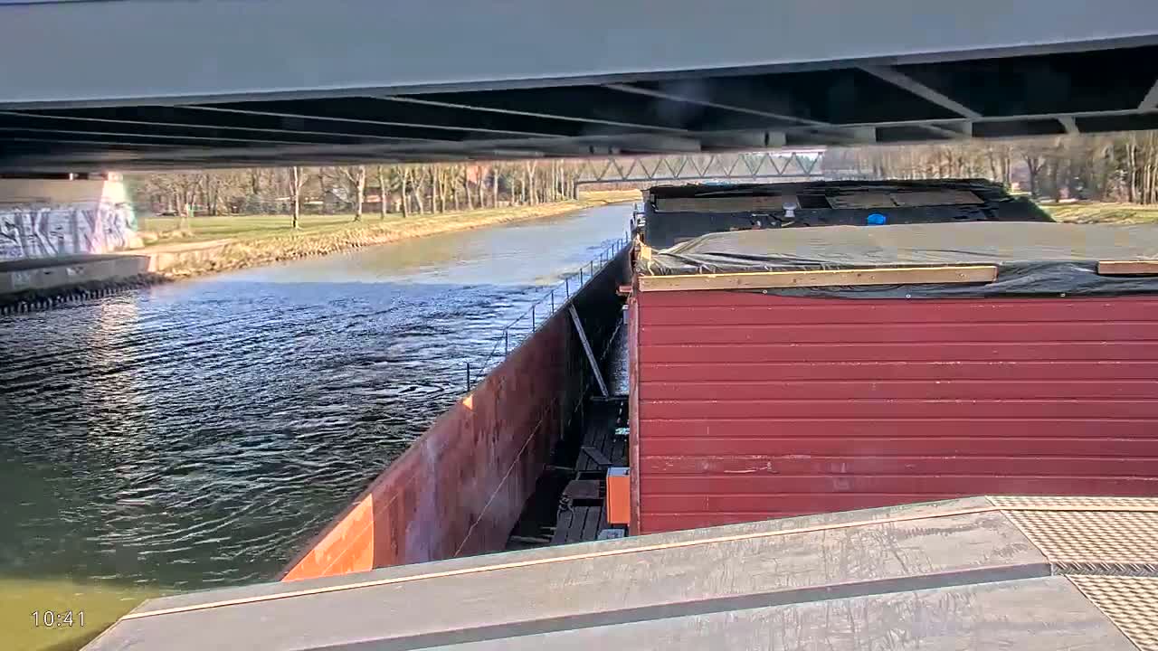 A red barge is traveling under a bridge on a calm canal, surrounded by trees and greenery.