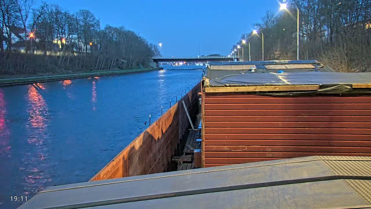 A barge travels down a canal at dusk under a clear, twilight sky, passing under a bridge with lights lining the banks.