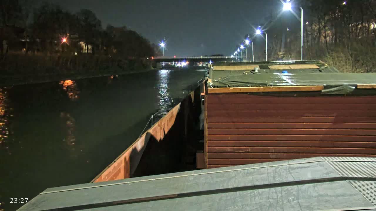 A nighttime view from aboard a barge on a canal, under artificial light, shows another barge alongside and a bridge in the distance.