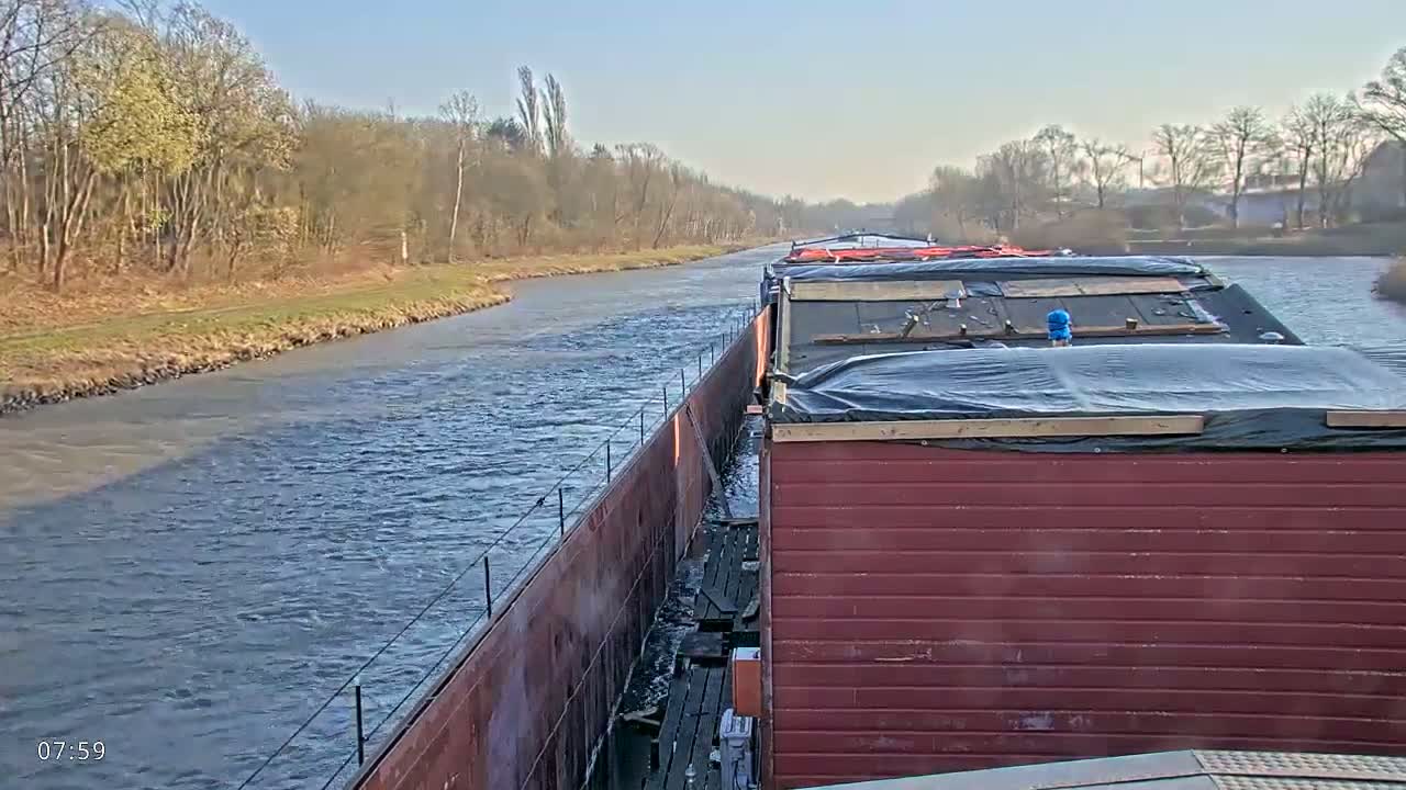 A barge travels down a calm river on a sunny day, past a wooded shoreline.