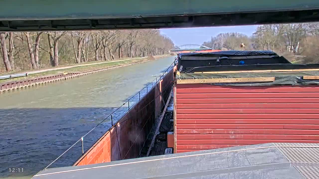 A barge travels down a canal on a sunny day, passing under a bridge and alongside a tree-lined bank.