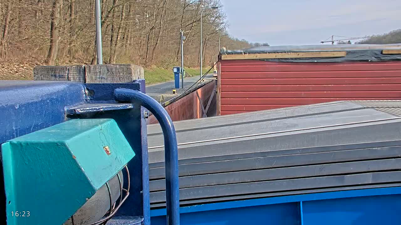 A barge carrying a stack of gray metal sheets is seen from a close, low angle, passing through a canal under a sunny sky near a wooded area.