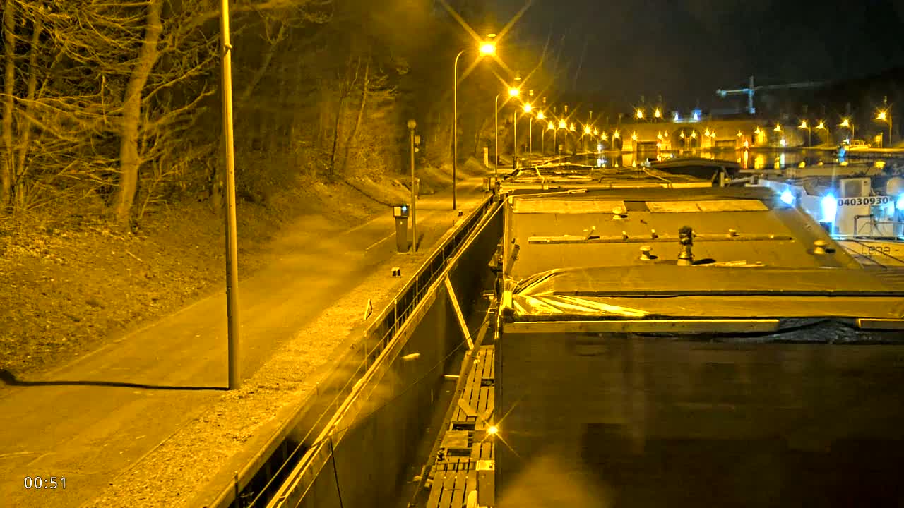 A nighttime view from a canal lock shows numerous barges lined up along the waterway, illuminated by bright streetlights lining a path beside bare trees.