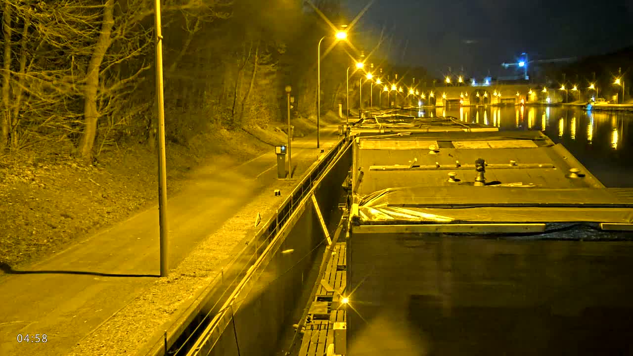At night, a series of barges are moored along a canal, illuminated by streetlights lining a path beside the water, with trees visible on the opposite bank.