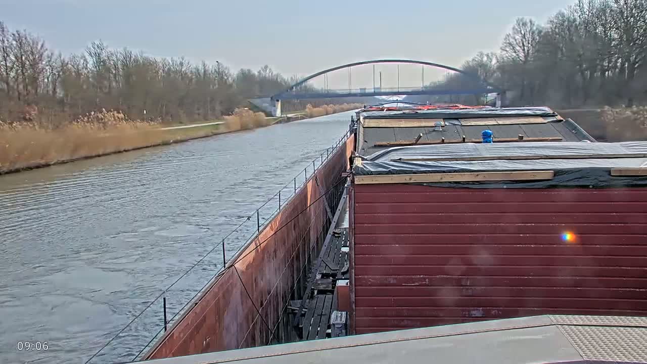 A barge travels down a canal on a partly cloudy day, passing under a bridge.
