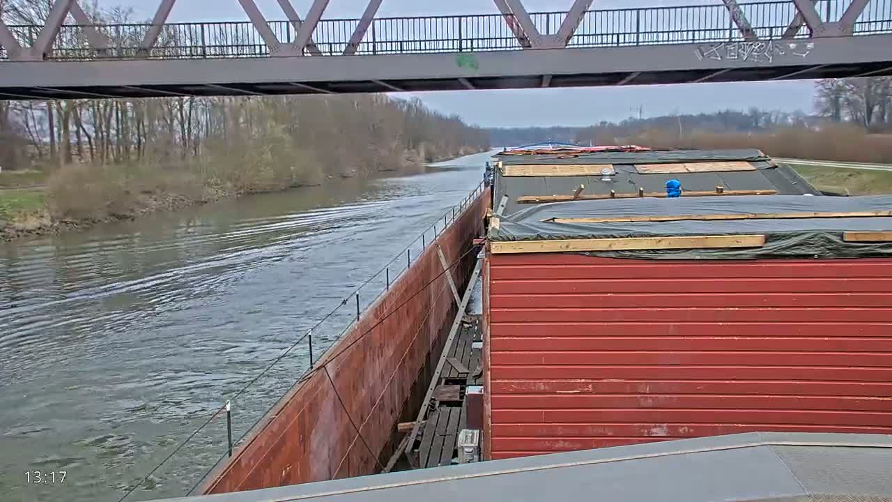 A barge with a red side and a rusty side travels under a bridge on a calm, overcast day.