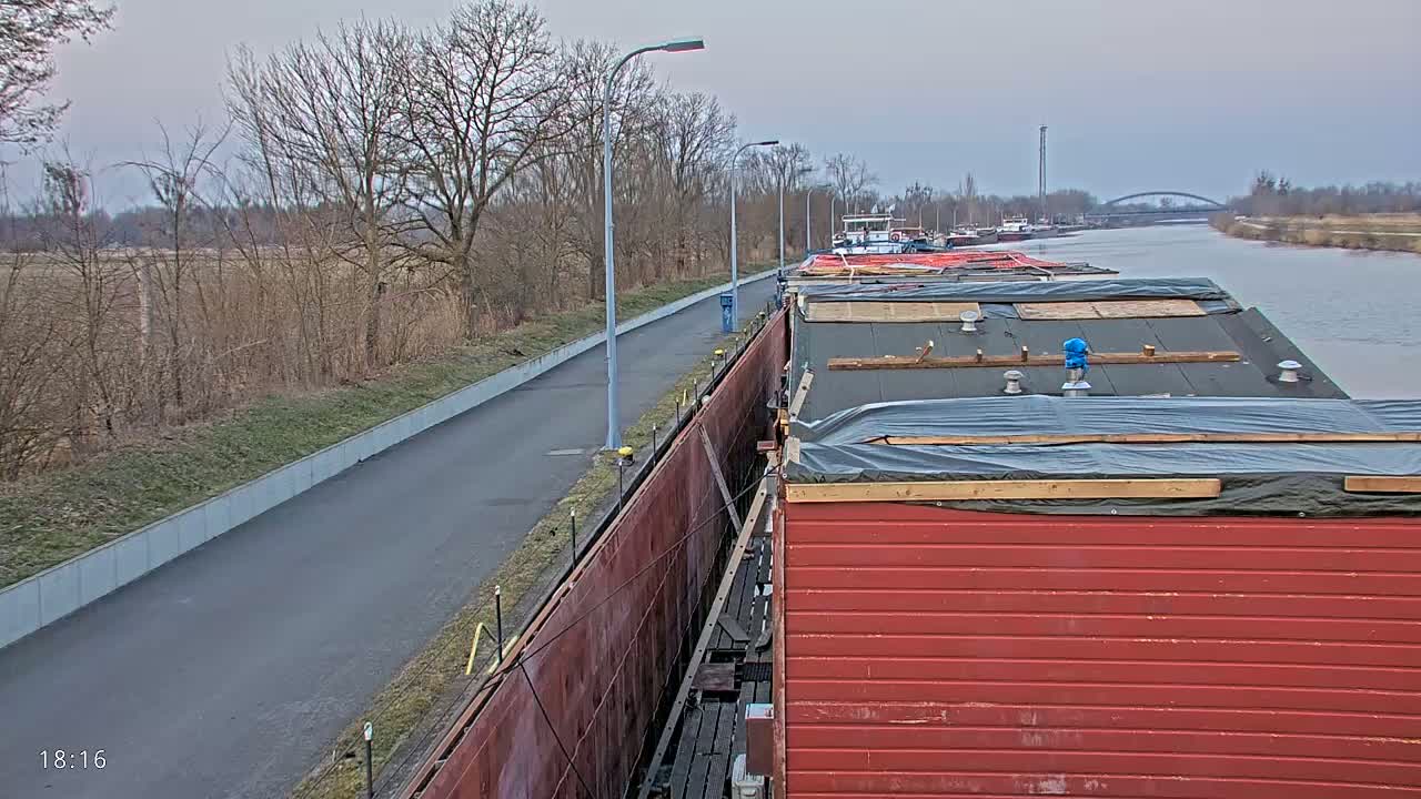 A paved path runs alongside a canal where several barges are moored, under an overcast sky.