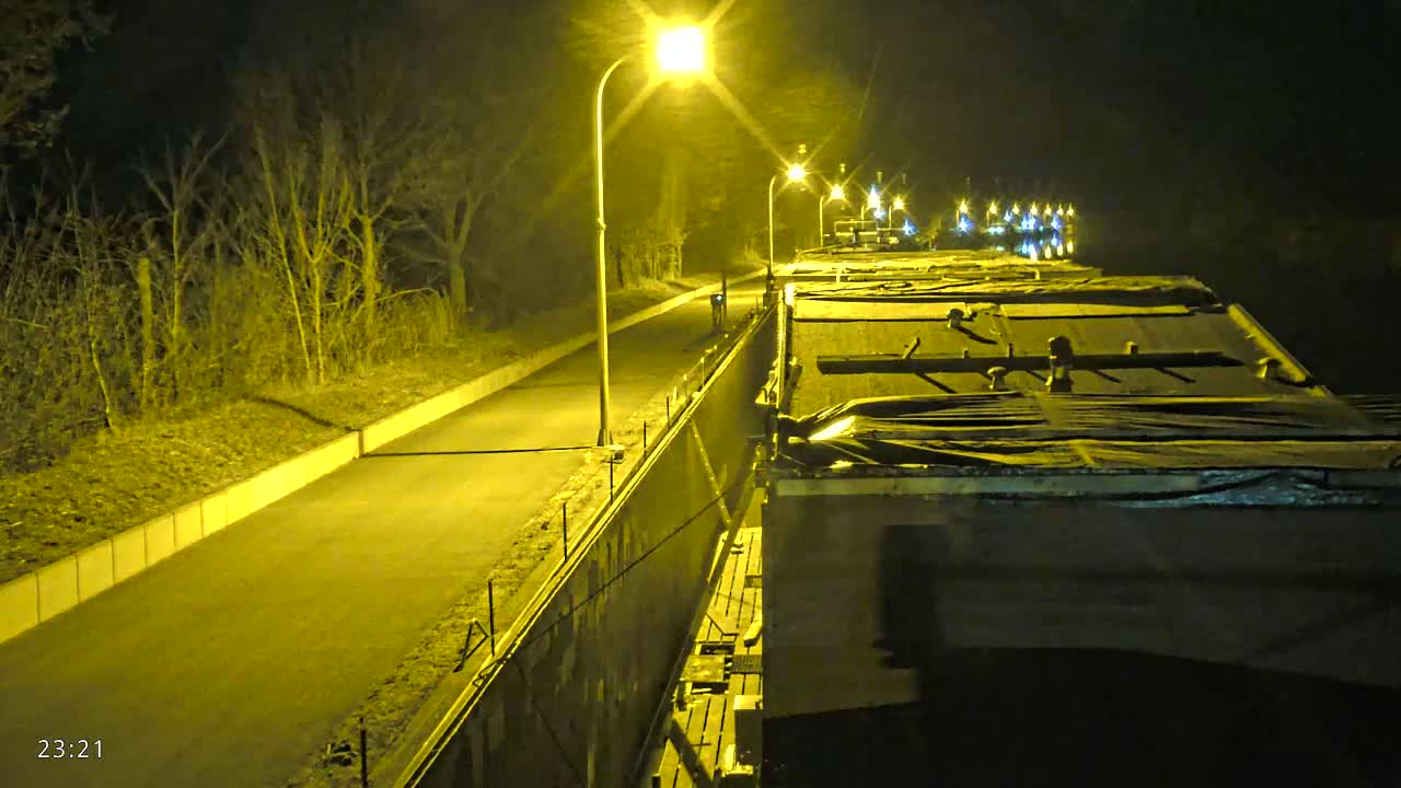 A paved road at night, illuminated by streetlights, runs alongside a canal where several covered barges are moored.