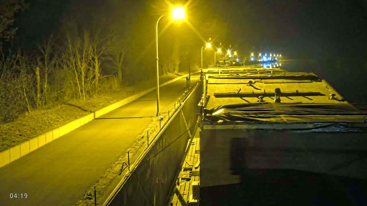 At night, a paved path next to a canal is illuminated by streetlights, showing several barges moored along the waterway.