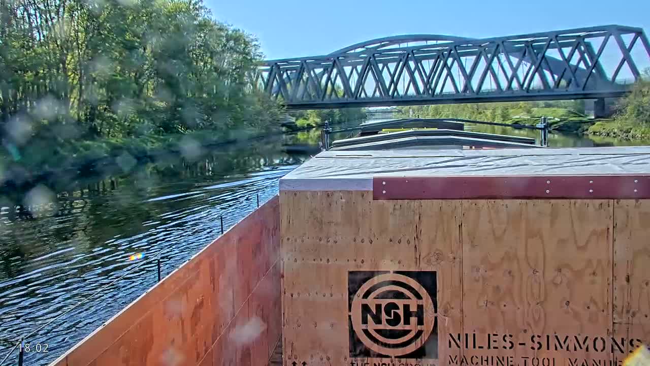 A barge carrying cargo travels on a calm river under a metal bridge on a sunny day.