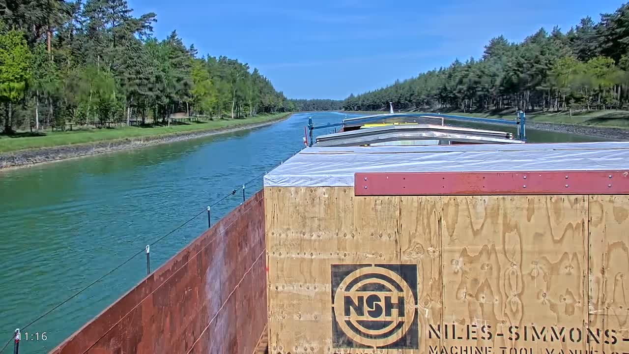 A barge travels down a canal on a sunny day, lined with lush green trees.