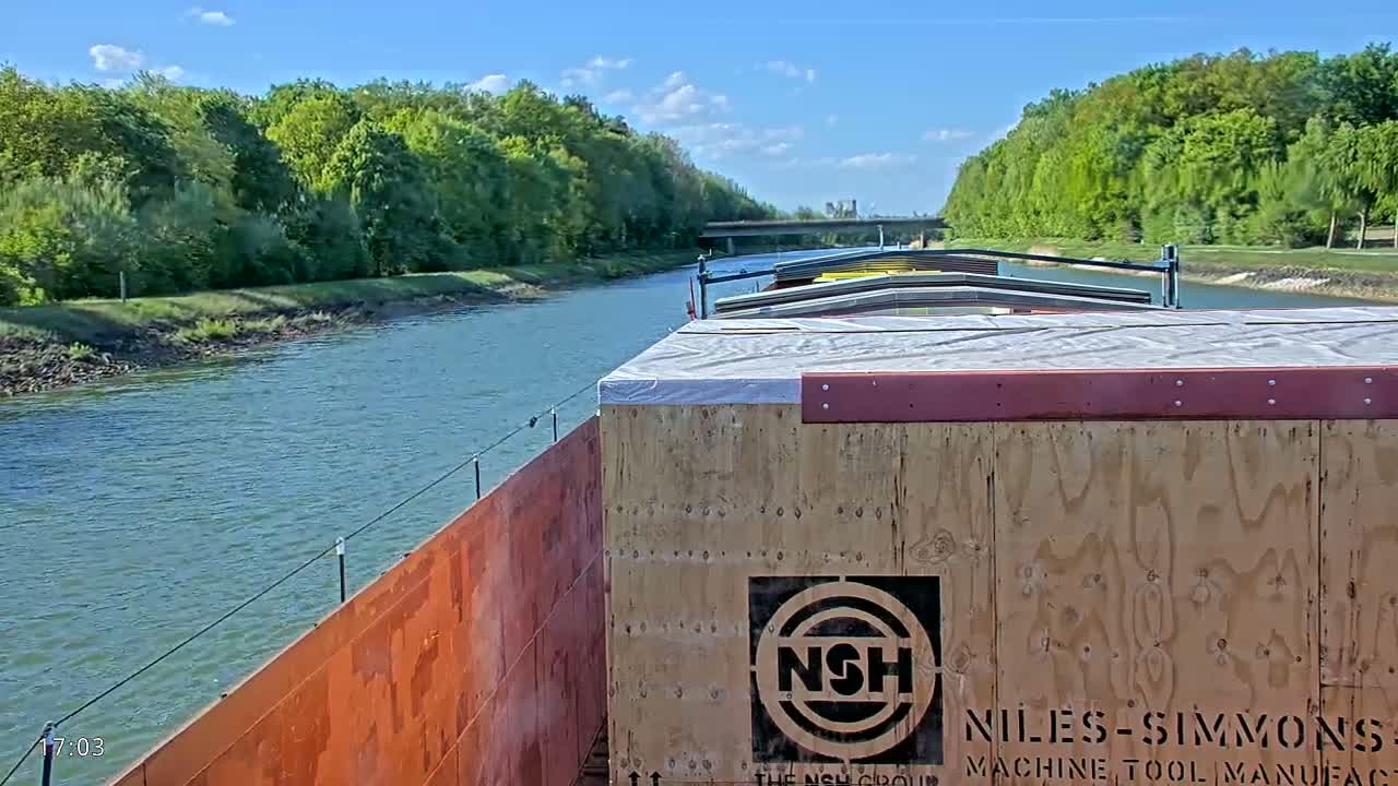 A barge travels down a canal on a sunny day, surrounded by lush green trees.
