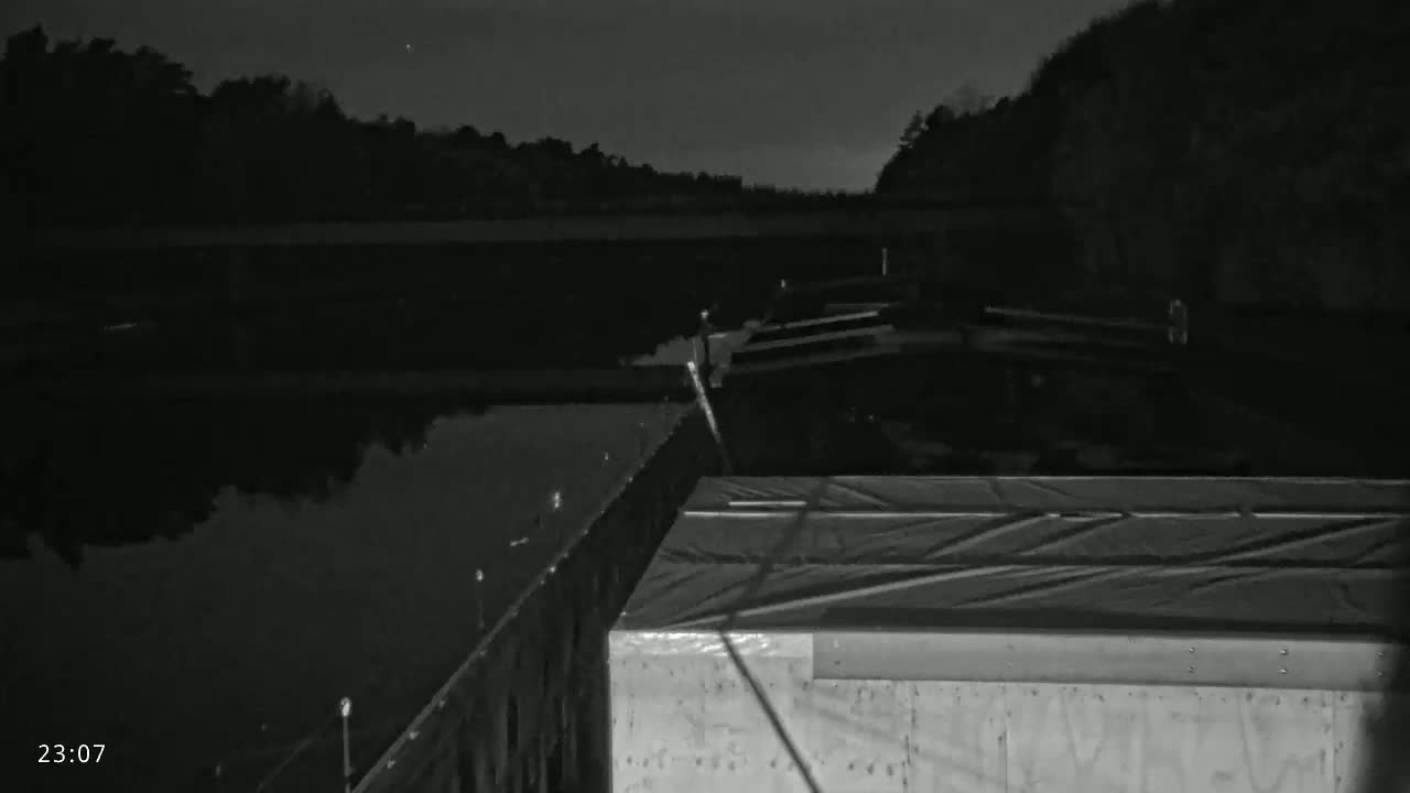 A nighttime, low-light image shows a dark body of water next to a structure covered in a tarp, with trees in the background under a clear, dark sky.