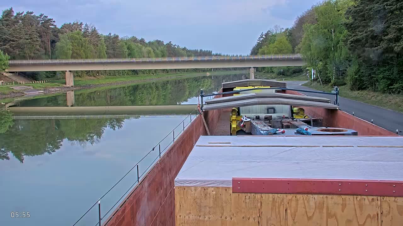 A barge carrying cargo travels along a canal under a bridge on an overcast day.