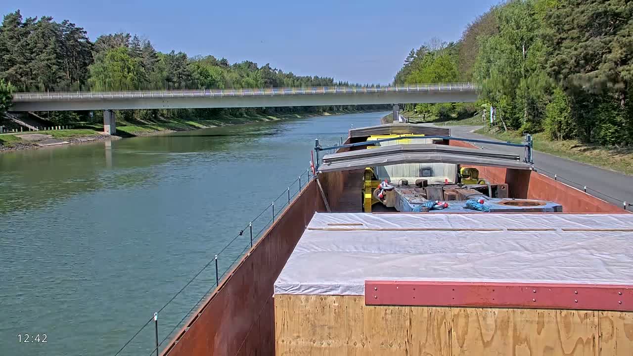 A barge carrying cargo travels on a calm river under a bridge on a sunny day, with lush green trees lining the banks.