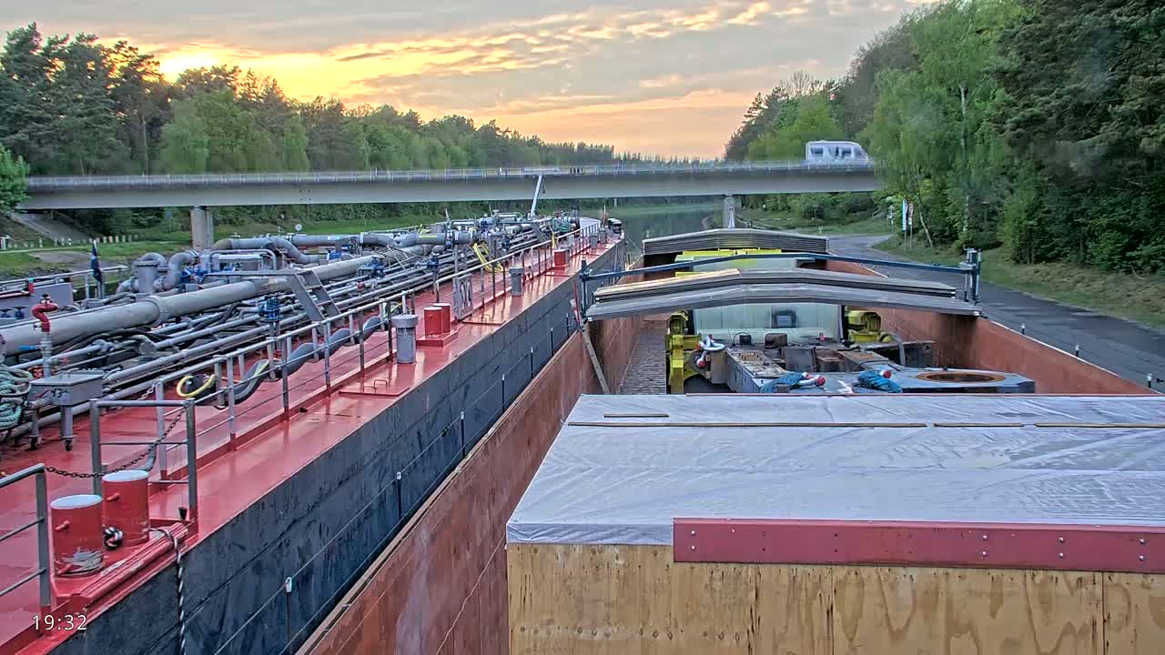 Two barges, one carrying piping and the other carrying covered cargo, are moored side-by-side in a canal at sunset under a mostly clear sky.