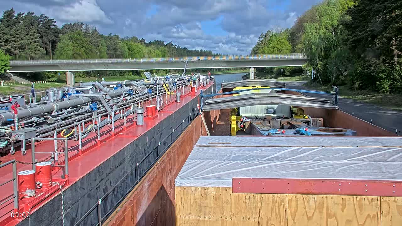 Two barges, one carrying pipes and the other carrying cargo covered in plastic sheeting, are traveling on a canal under a bridge on a partly cloudy day.