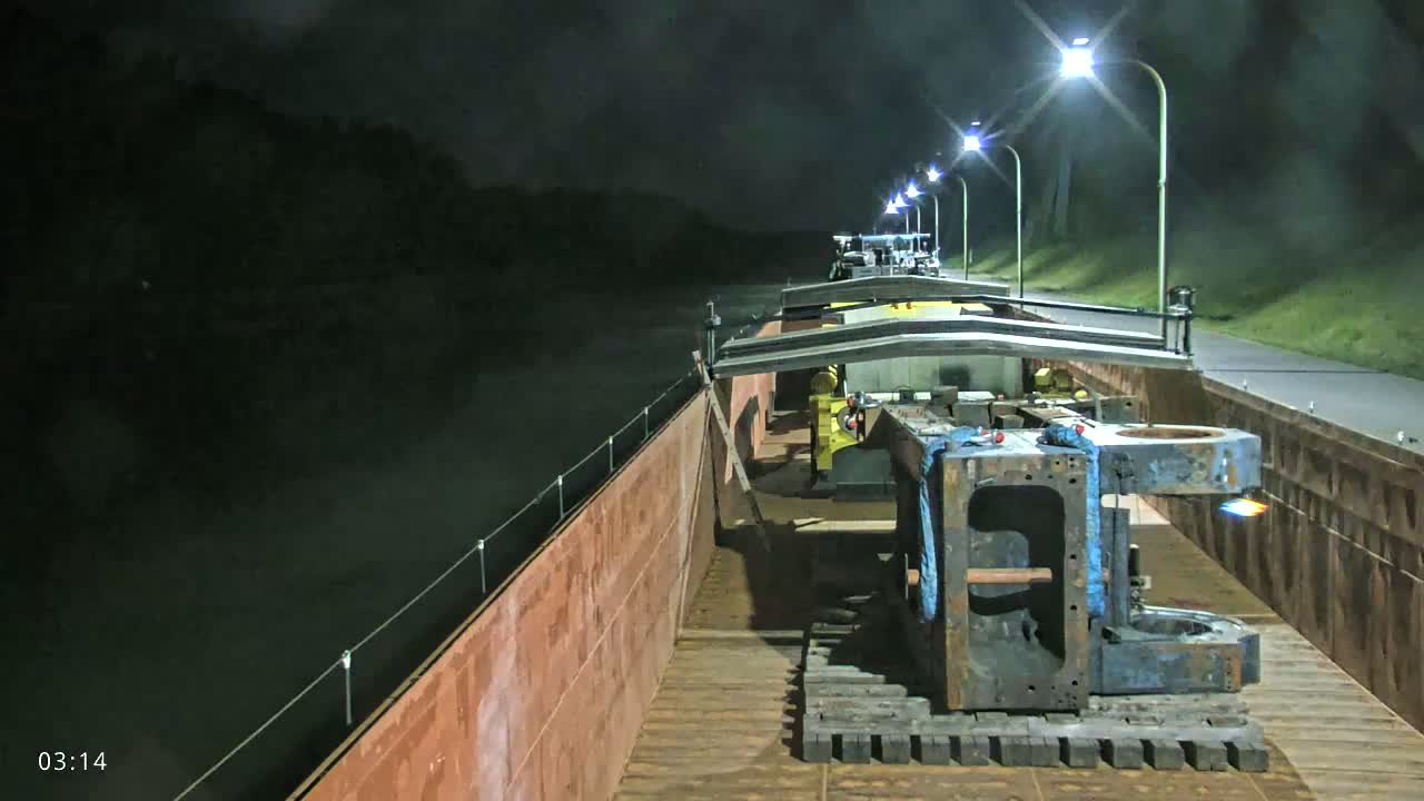 A barge carrying large pieces of industrial machinery is lit by lamps at night on a calm body of water.