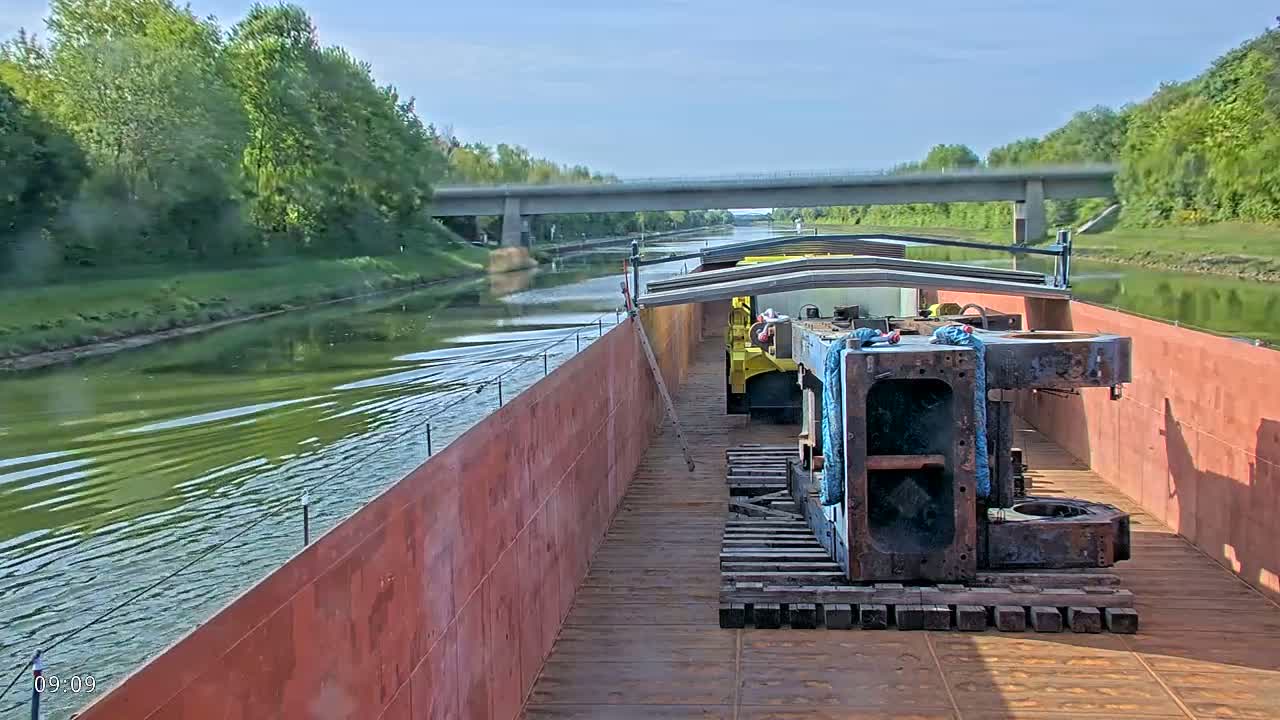 A barge carrying large metal objects travels down a canal on a sunny day, passing under a bridge lined with green trees.