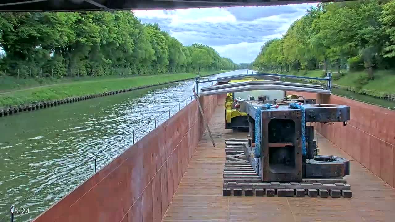 A barge carrying large metal equipment travels down a canal lined with lush green trees under a partly cloudy sky.