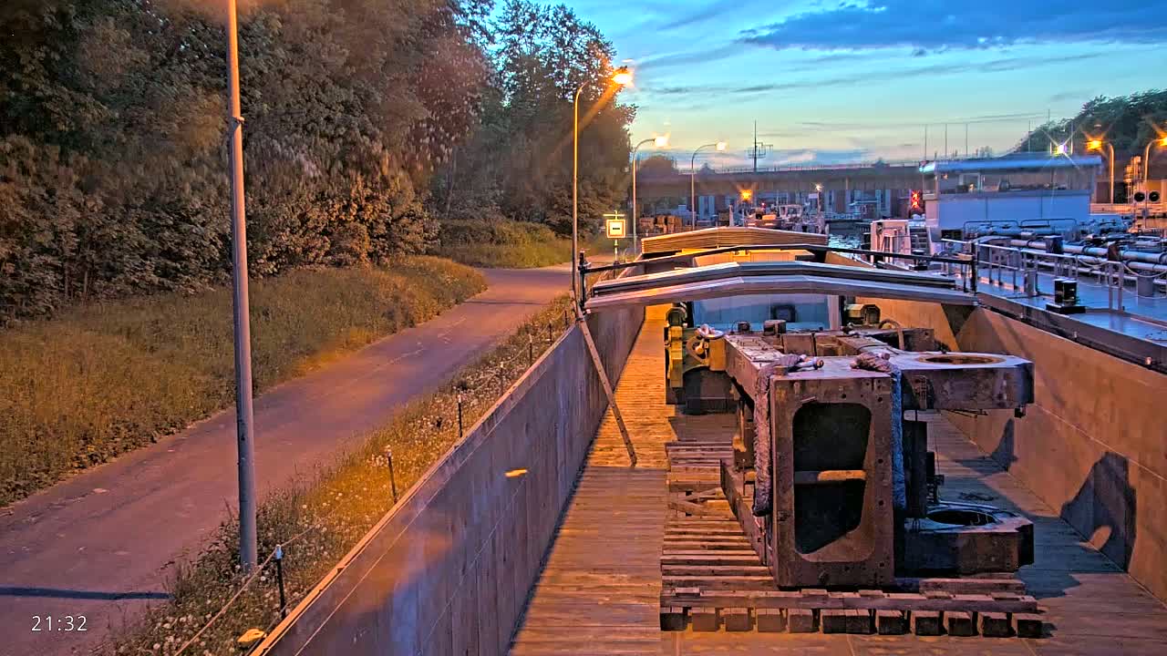 Large pieces of industrial machinery are secured on a barge at dusk, alongside a paved path and lush green vegetation.