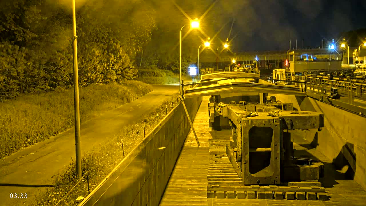 A large piece of industrial machinery sits on a barge at night, illuminated by several streetlights, beside a road lined with trees.