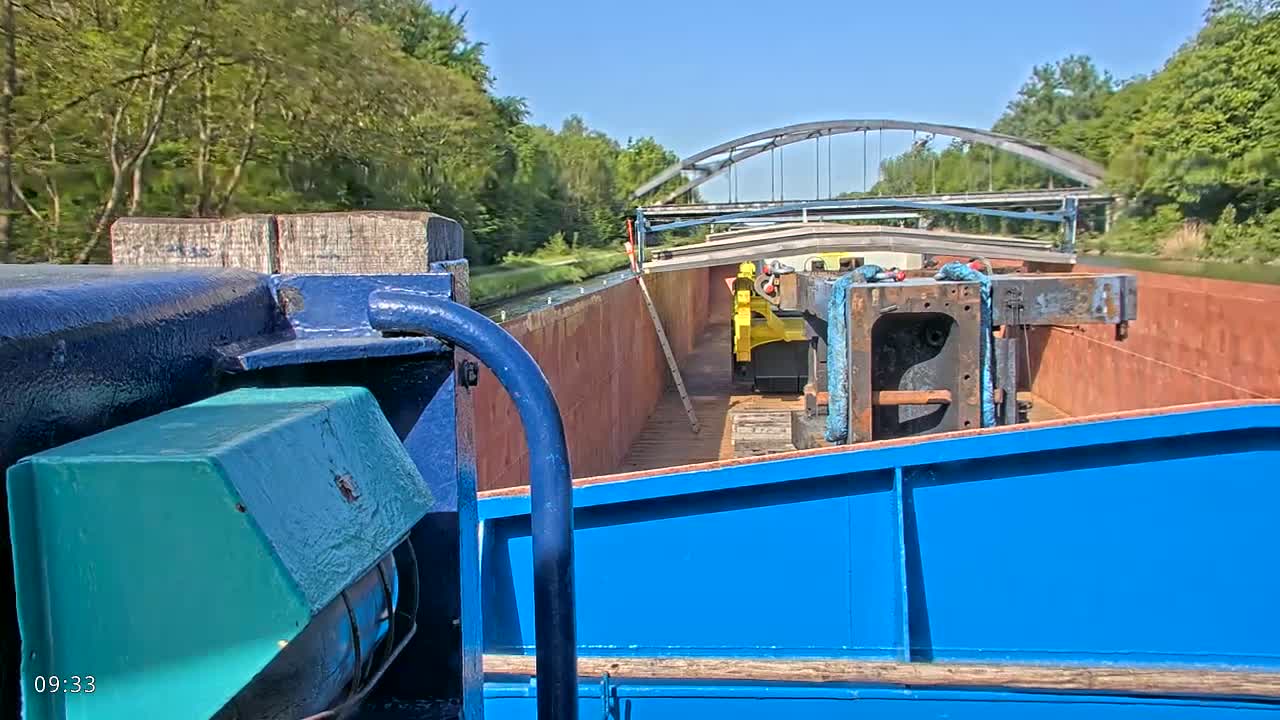 A barge carrying large pieces of machinery travels on a canal under a metal bridge on a sunny day.