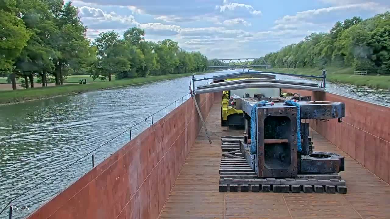 A barge carrying large pieces of machinery travels down a canal under a partly cloudy sky, lined with lush green trees.