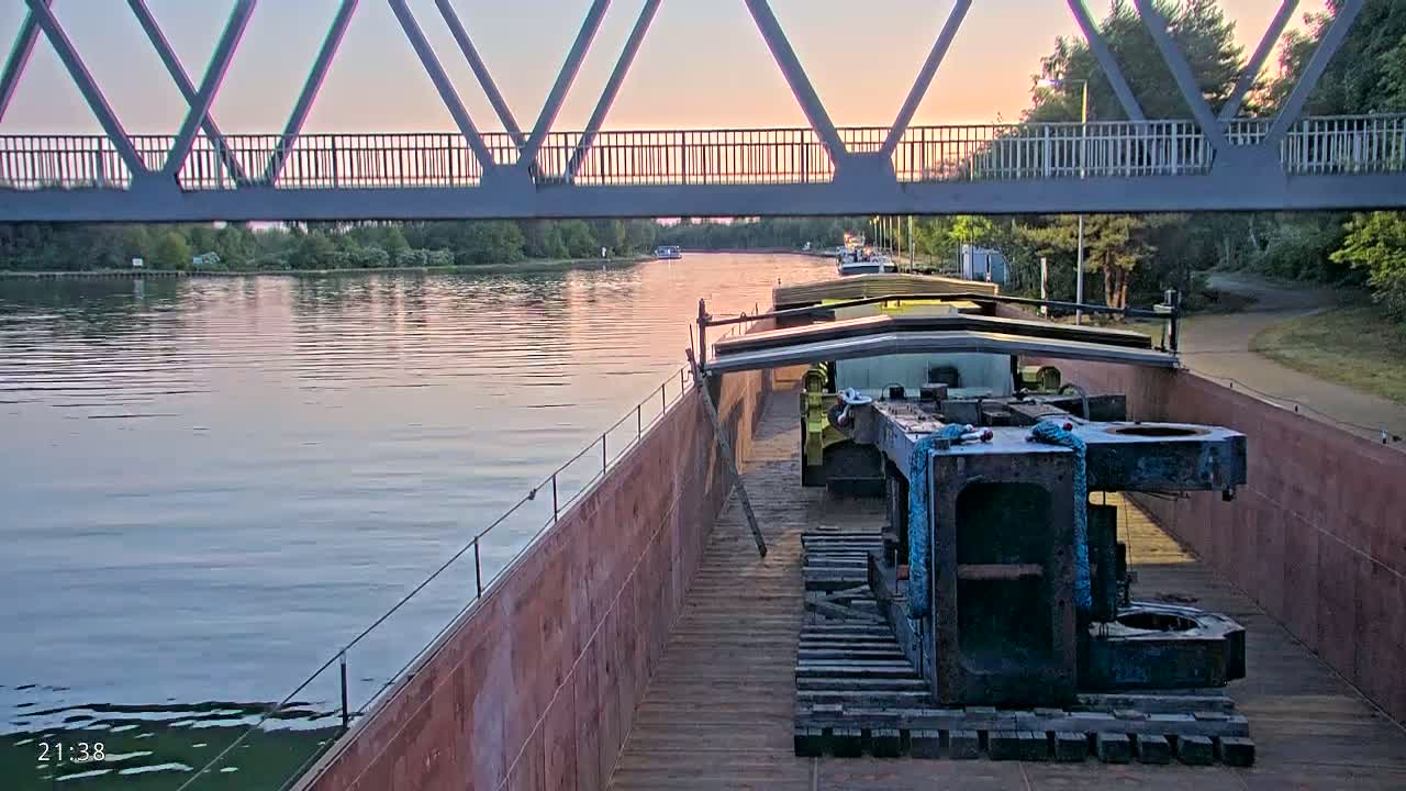 A barge carrying large pieces of industrial machinery travels on a calm river at sunset under a pedestrian bridge.