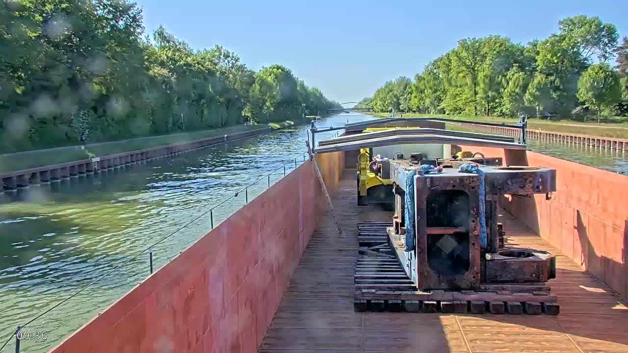 A barge carrying large metal components travels down a canal on a sunny day.