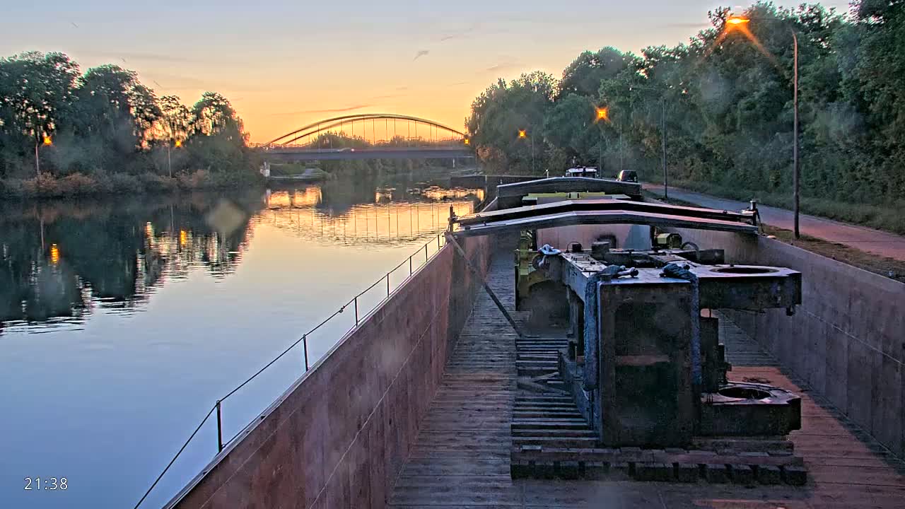 A barge carrying large pieces of machinery is moored on a calm river at sunset, with a bridge visible in the distance.
