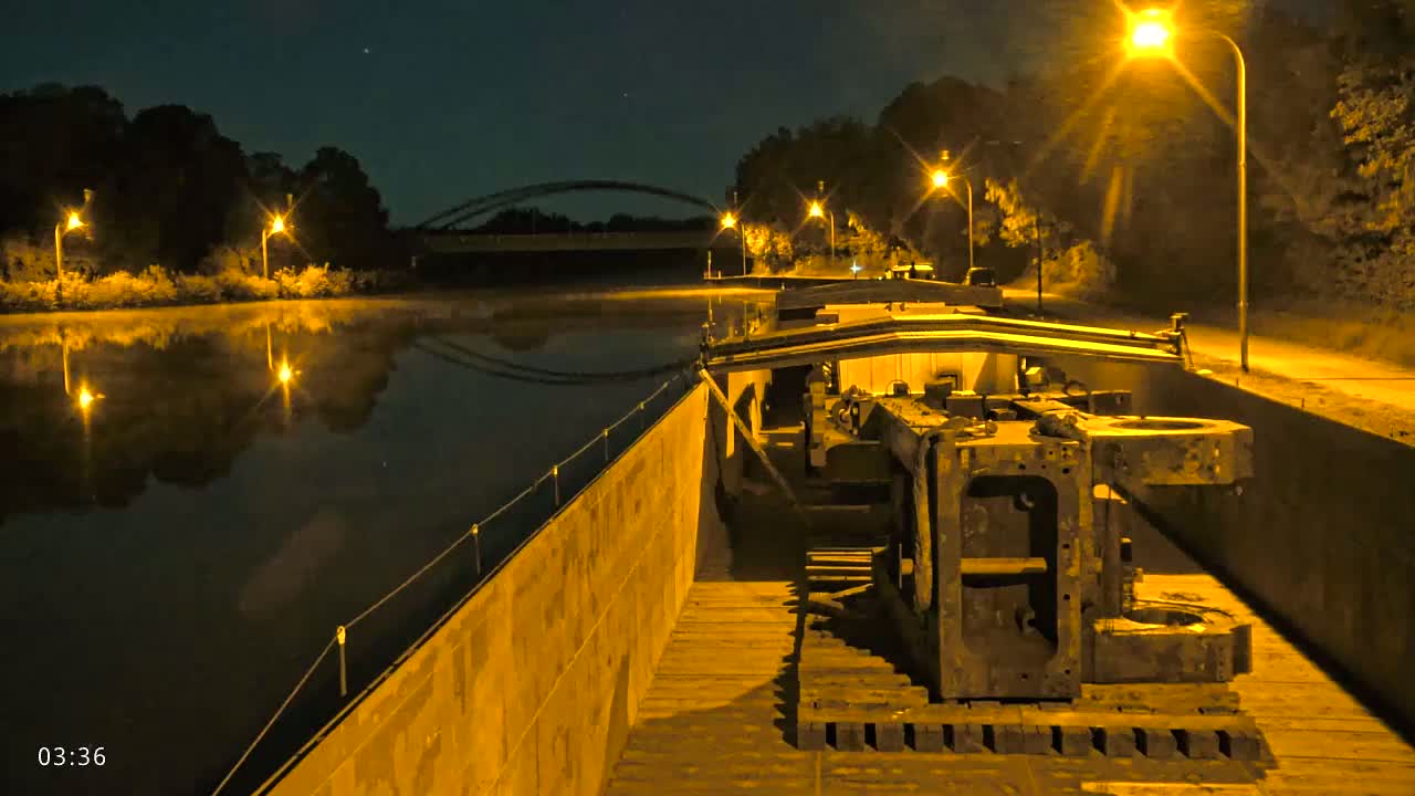 A barge carrying large pieces of machinery is moored at night next to a calm body of water under illuminated streetlights.