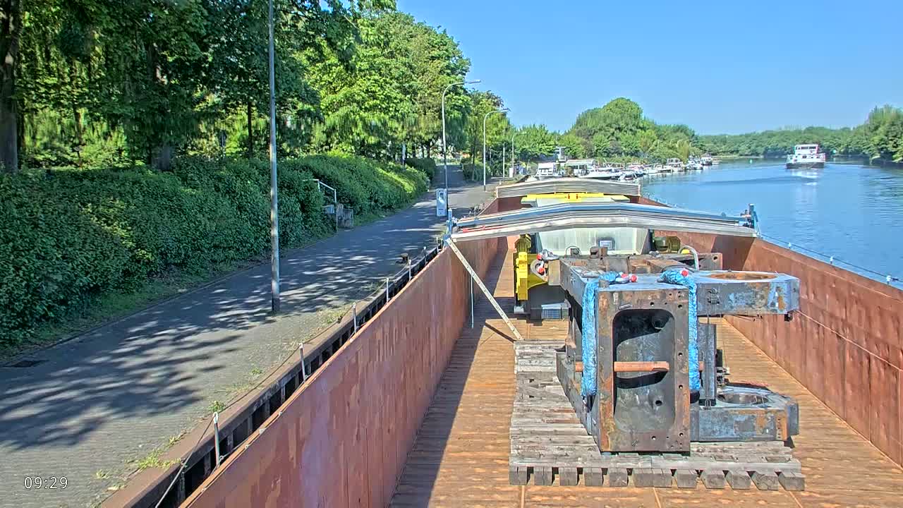 A barge carrying large pieces of machinery is moored alongside a paved walkway that runs next to a riverbank lined with lush green vegetation under a sunny sky.