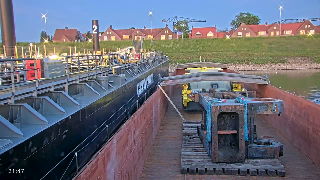 Two barges are moored together at dusk, one carrying industrial equipment on wooden pallets, while the other appears to be a fuel barge, with houses visible in the background.