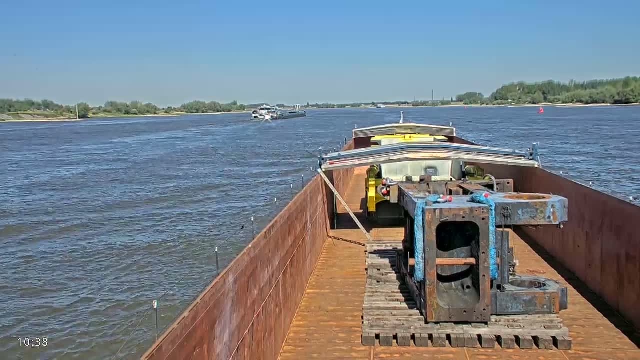 A barge carrying heavy machinery travels down a river on a sunny day.