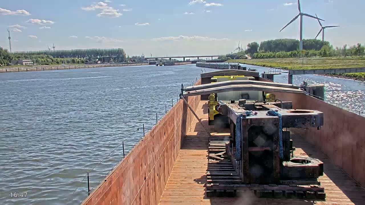 A barge carrying large pieces of industrial equipment travels down a waterway on a sunny day under a partly cloudy sky, with wind turbines visible in the distance.