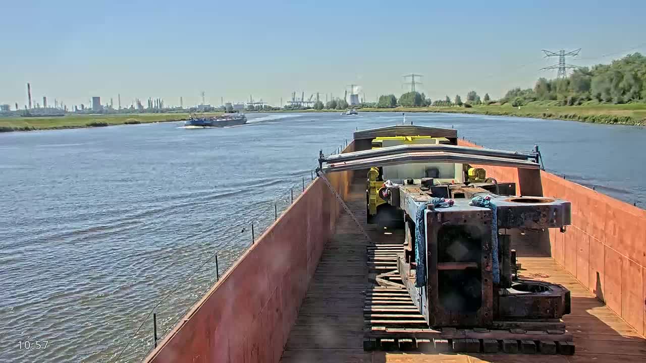 A barge carrying large pieces of industrial equipment travels down a wide river on a sunny day, with an industrial landscape visible in the distance.