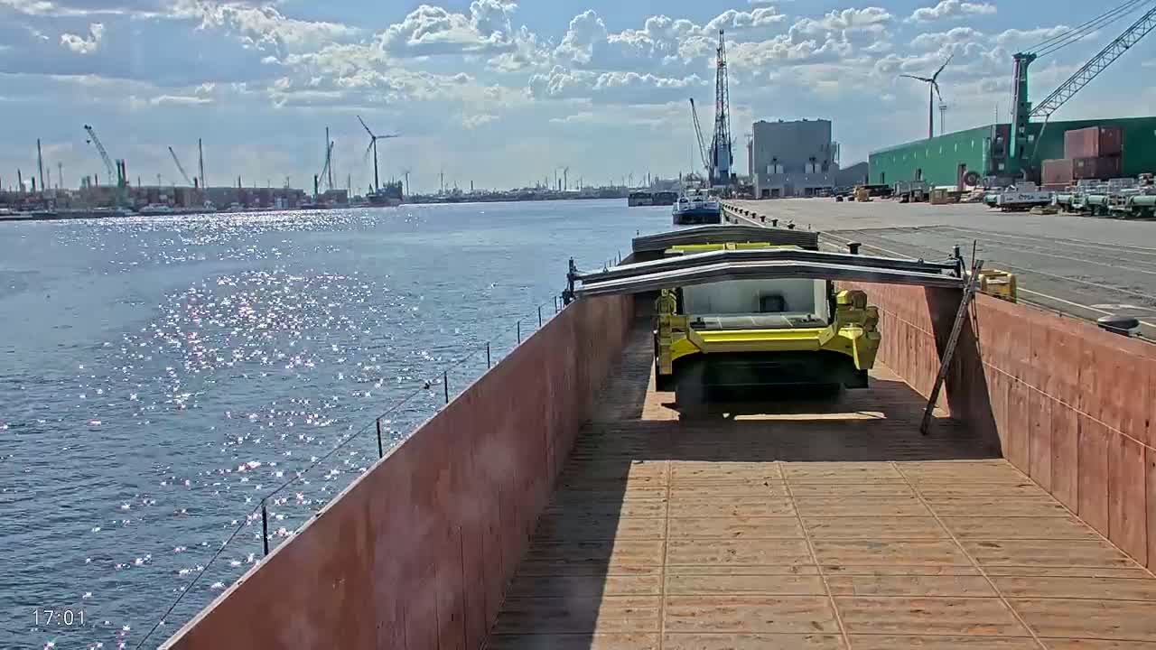 A barge carrying yellow machinery is on a waterway under a partly cloudy sky.