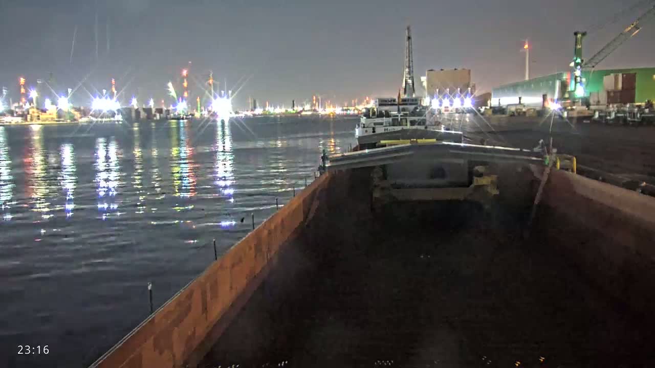 At night, from the interior of a barge, a calm body of water reflects the lights of a distant industrial port.
