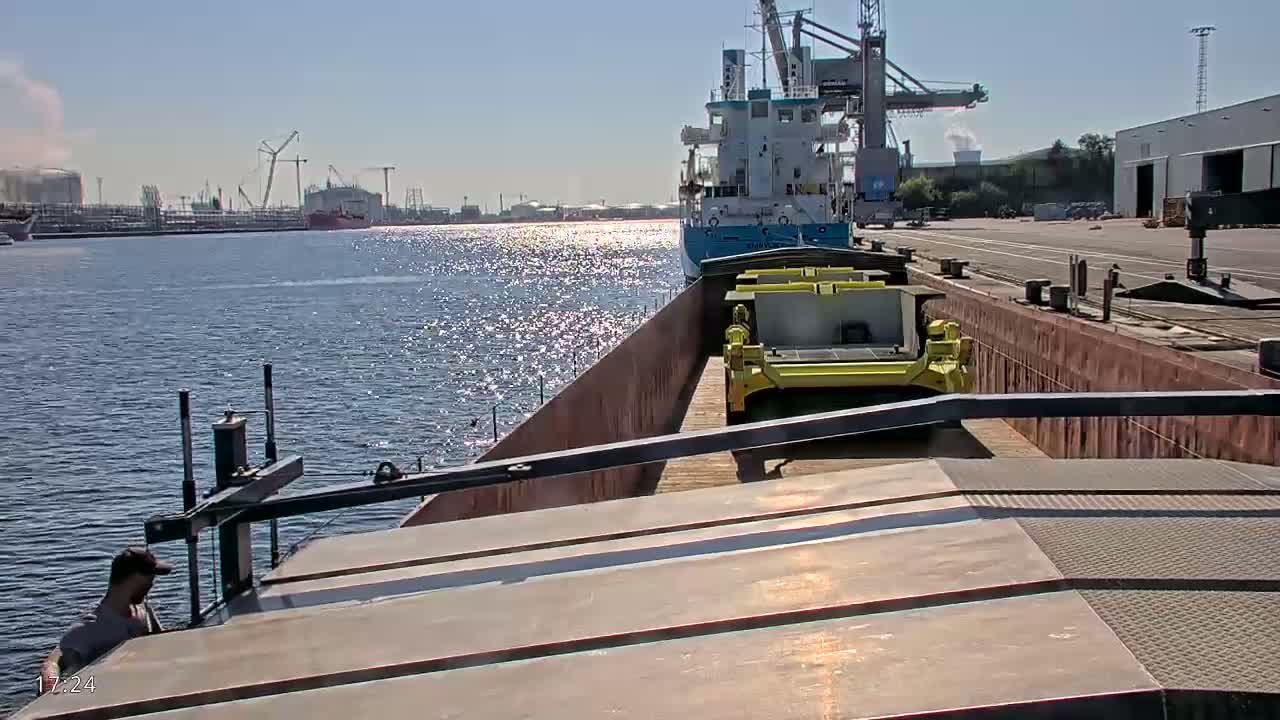 A barge carrying yellow machinery is moored alongside a larger ship in a sunny harbor.