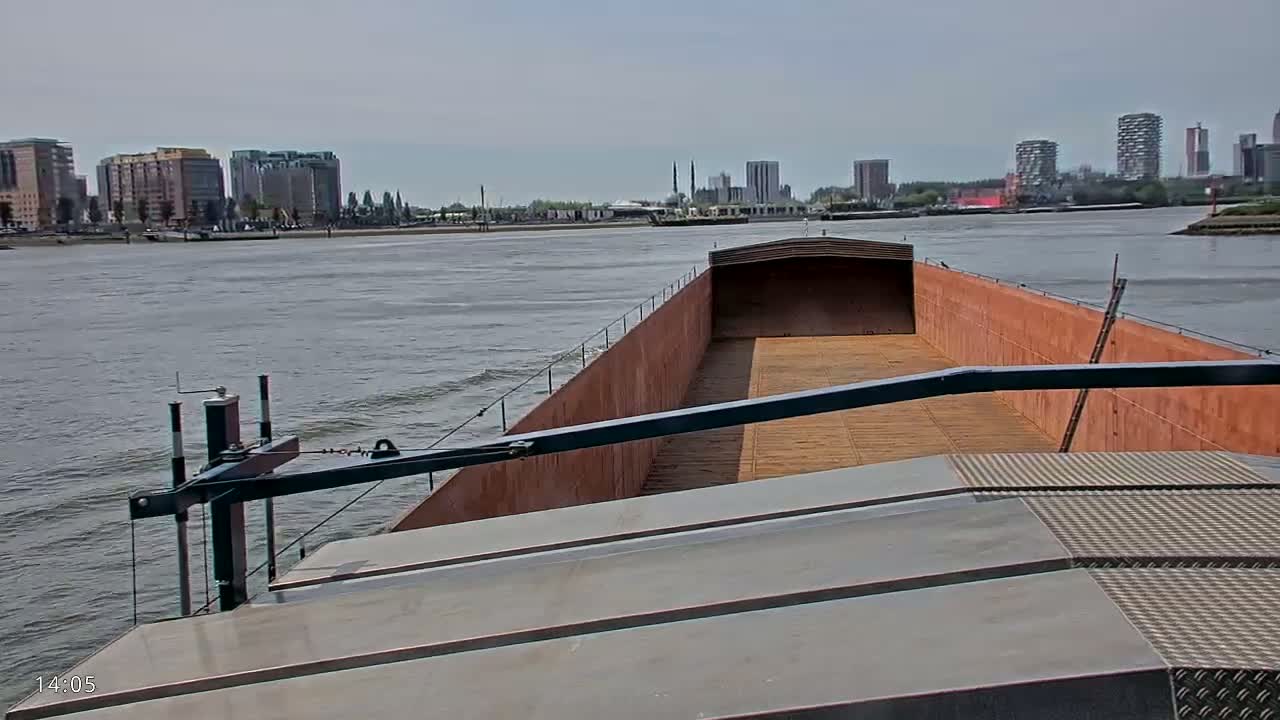A barge travels on a river under a cloudy sky past a city skyline.