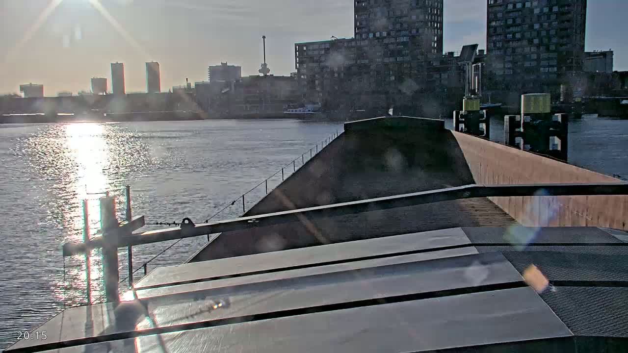 A barge's deck is shown, with a city skyline and calm, sunlit water visible in the background on a bright day.