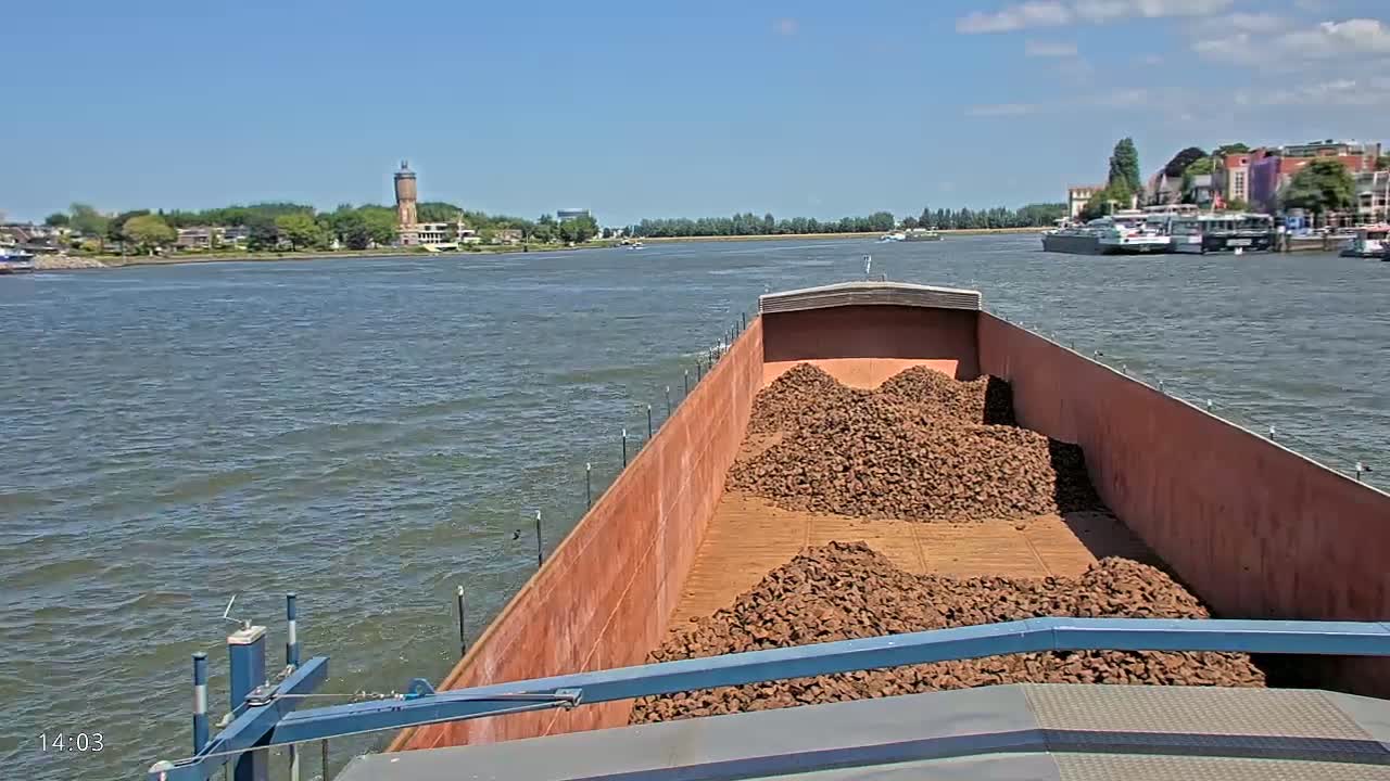 A barge filled with piles of dark brown material travels on a waterway under a sunny sky.