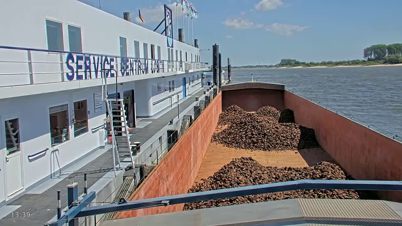 A barge loaded with dark brown rocks is docked alongside a white building on a sunny day next to a river.