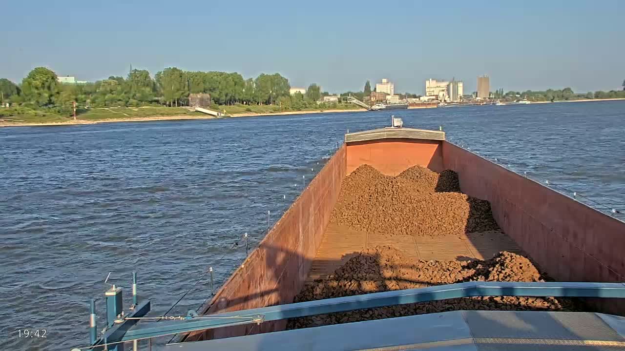 A barge filled with a large pile of reddish-brown rocks travels down a river on a sunny day.