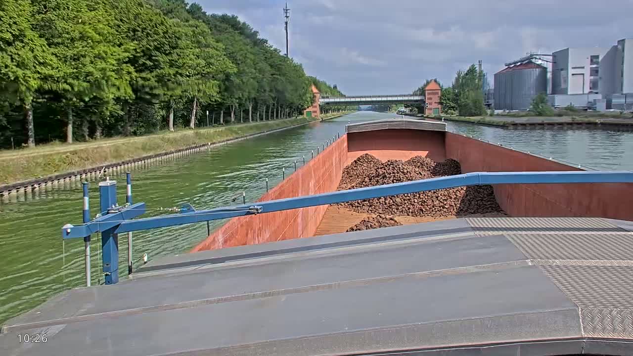 A barge loaded with a large pile of dark-brown material travels down a canal on a sunny day, lined with green trees and industrial buildings in the distance.