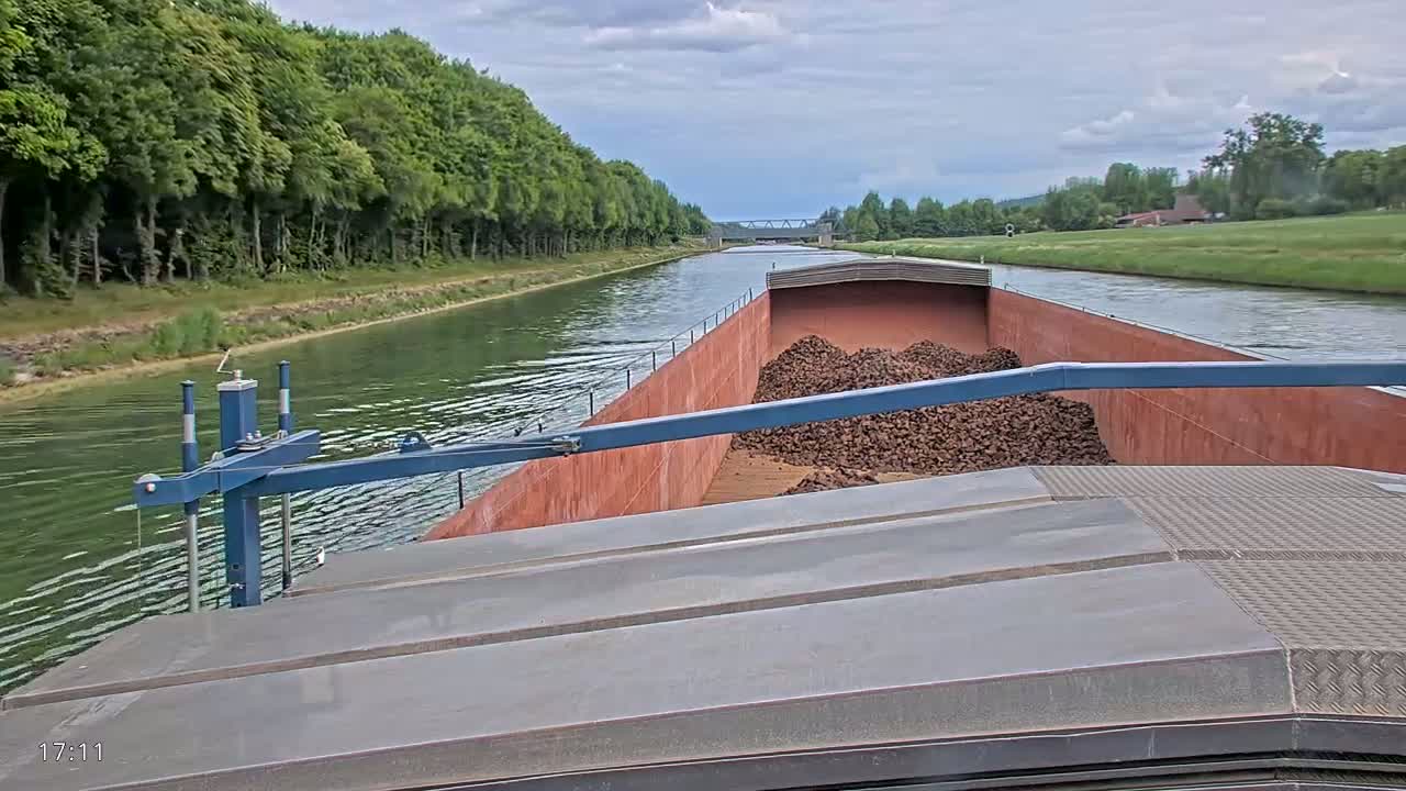 A barge carrying a large pile of dark-brown material travels down a canal lined with green trees under a cloudy sky.