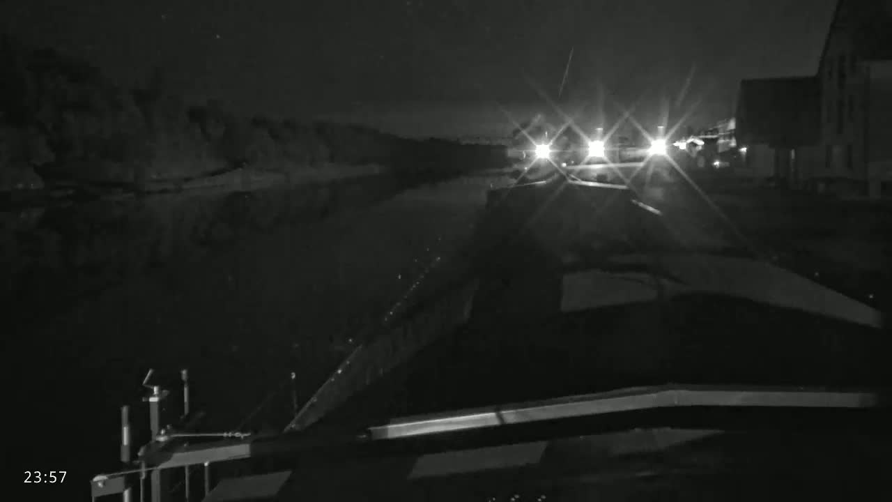 A nighttime view from a boat on a calm waterway, showing approaching headlights and a dark shoreline.